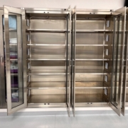 Empty stainless steel storage cabinet with glass double doors, standing on a smooth gray floor against a light beige wall. The cabinet features multiple adjustable shelves and a clean, modern design with a brushed metal finish