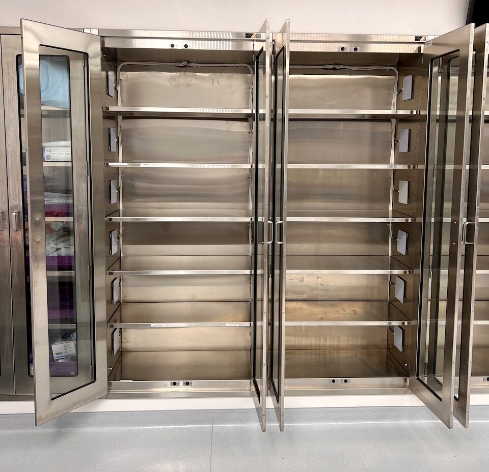 Empty stainless steel storage cabinet with glass double doors, standing on a smooth gray floor against a light beige wall. The cabinet features multiple adjustable shelves and a clean, modern design with a brushed metal finish